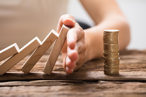A picture of a hand stopping wooden dominoes falling onto a pile of coins